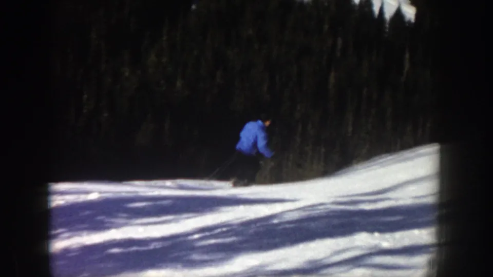 1961 Aspen home-movie frame showing a skier in a blue jacket moving across a snow-covered slope with evergreen forest behind.