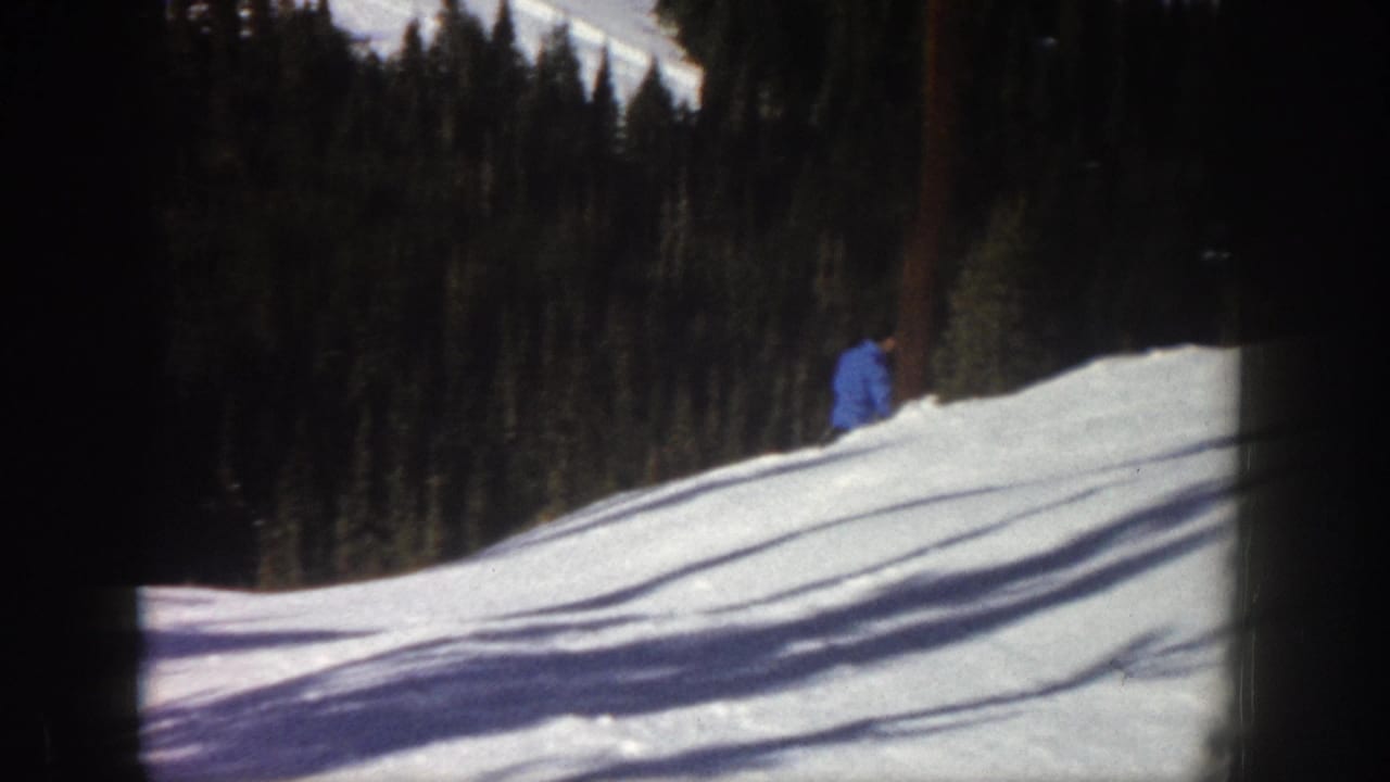 1961 Aspen 8mm frame with the skier partly distant on the right edge of a sunlit snowy slope below pine trees.