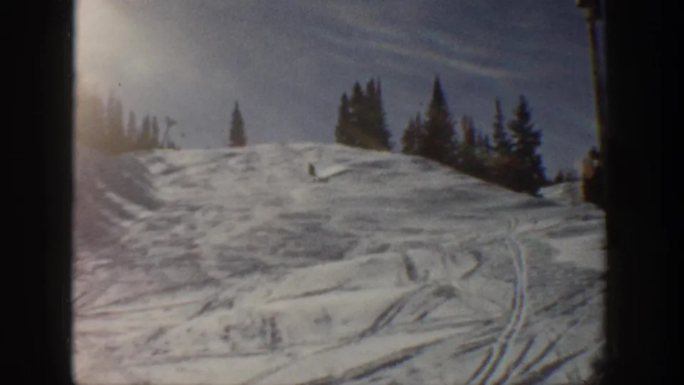 1961 Aspen 8mm frame showing a wider snow-covered hillside with ski tracks, small trees, and bright winter light.