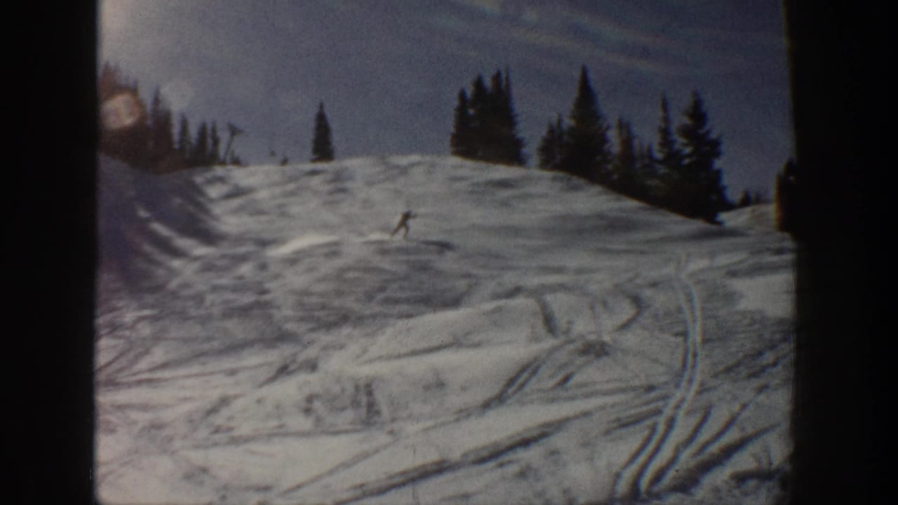 A lone skier is visible mid-slope on 1961 Aspen home-movie footage, with crisscrossed ski tracks and dark pines behind.