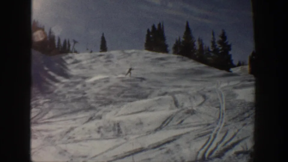 A lone skier is visible mid-slope on 1961 Aspen home-movie footage, with crisscrossed ski tracks and dark pines behind.