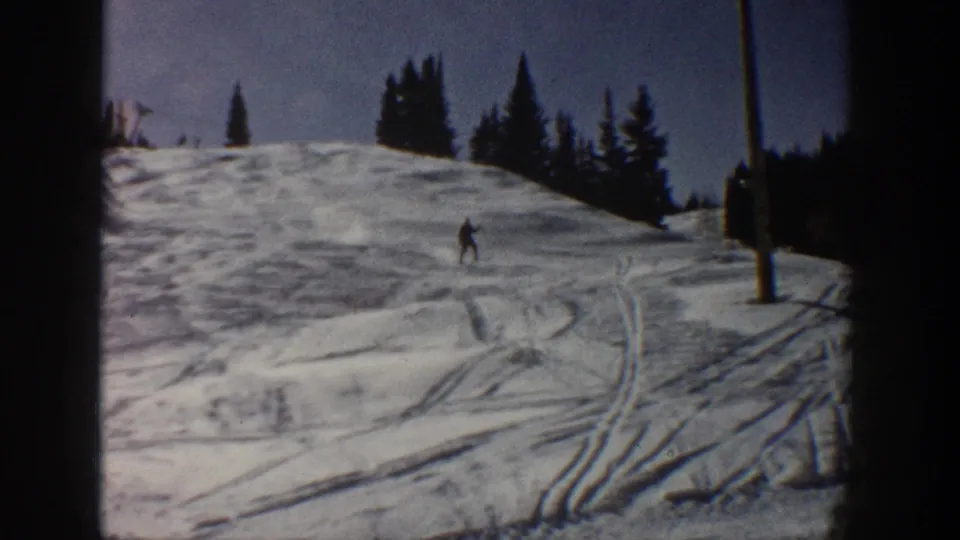 The skier appears lower on the Aspen slope, passing near a vertical pole with pines, blue sky, and marked snow visible around them.