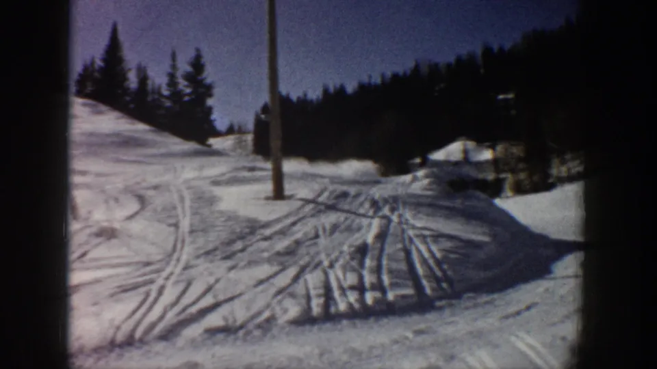 1961 Aspen home-movie frame showing a snowy slope marked by ski tracks, with evergreens and a wooden pole under blue sky.
