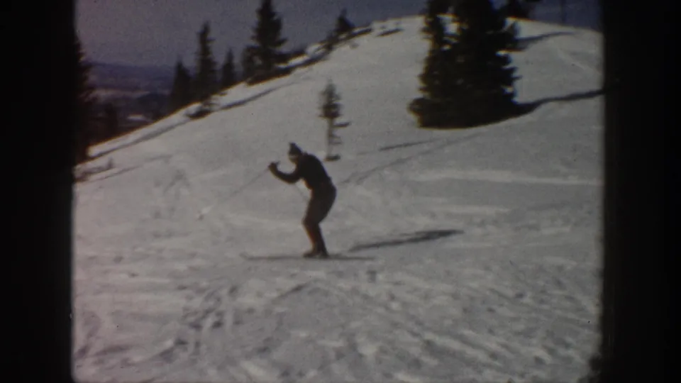 Lone skier in dark winter clothing descends a snowy Aspen hillside, with evergreens and ski tracks behind them.