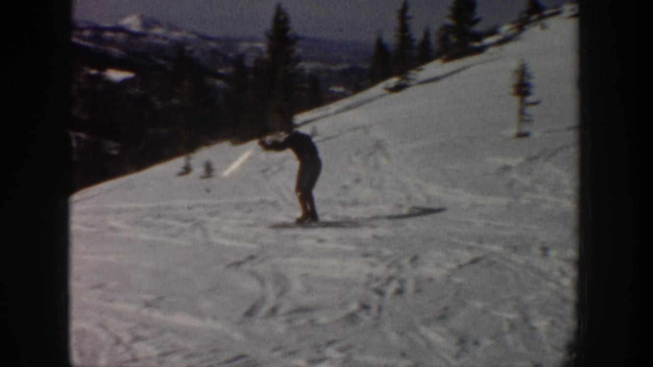 1961 Aspen home-movie frame showing a distant skier leaning into a snowy mountain slope with trees and ridges in the background.