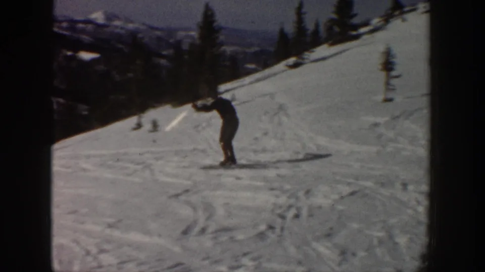 1961 Aspen home-movie frame showing a distant skier leaning into a snowy mountain slope with trees and ridges in the background.