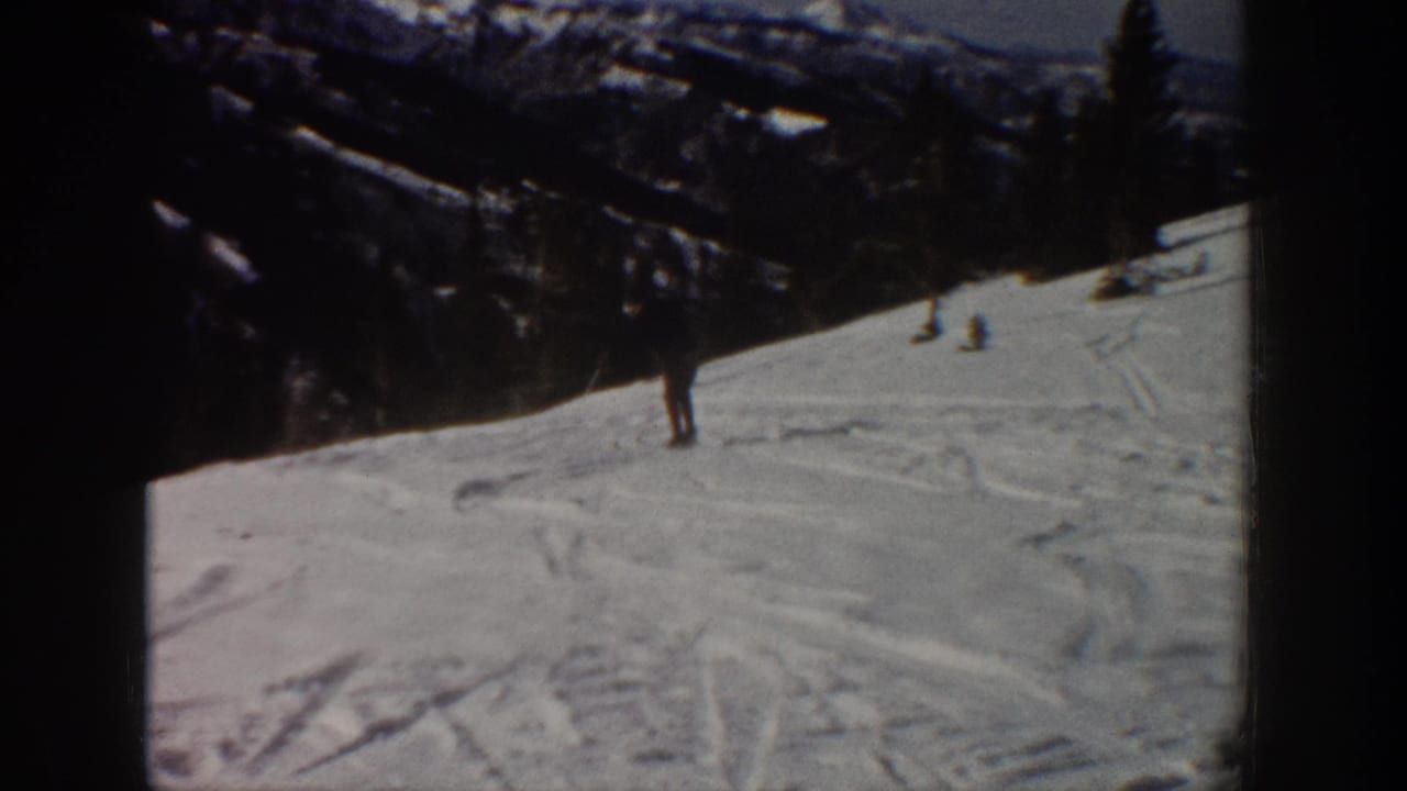 Distant skier on an Aspen snowy slope, framed by dark evergreen-covered mountains and visible 8mm film borders.