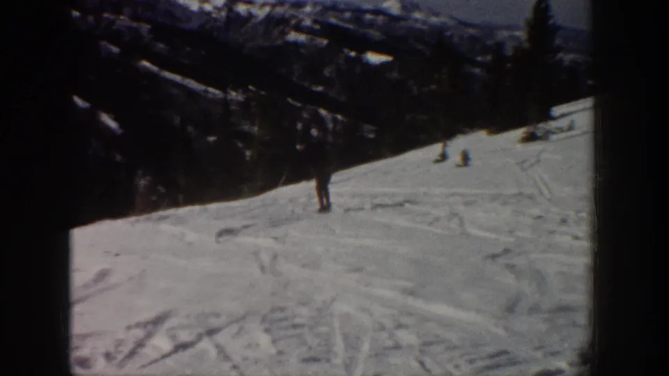 Distant skier on an Aspen snowy slope, framed by dark evergreen-covered mountains and visible 8mm film borders.