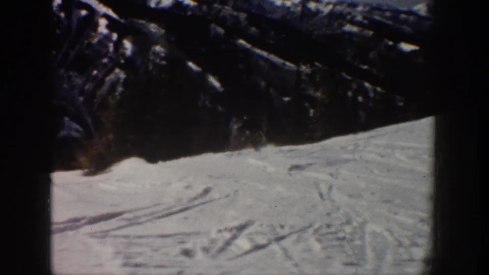 Tracked snowfield in the Aspen ski scene, with dark ridges and trees above and a faint skier presence near the slope edge.