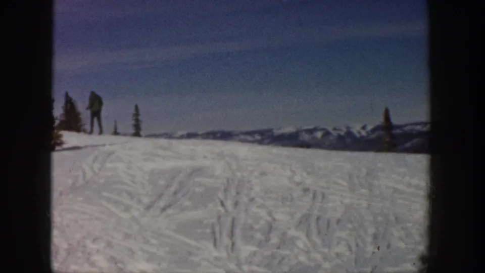 Wide Aspen ski slope in 1961 with small bundled figures, blue sky, evergreens, and distant snowy peaks.