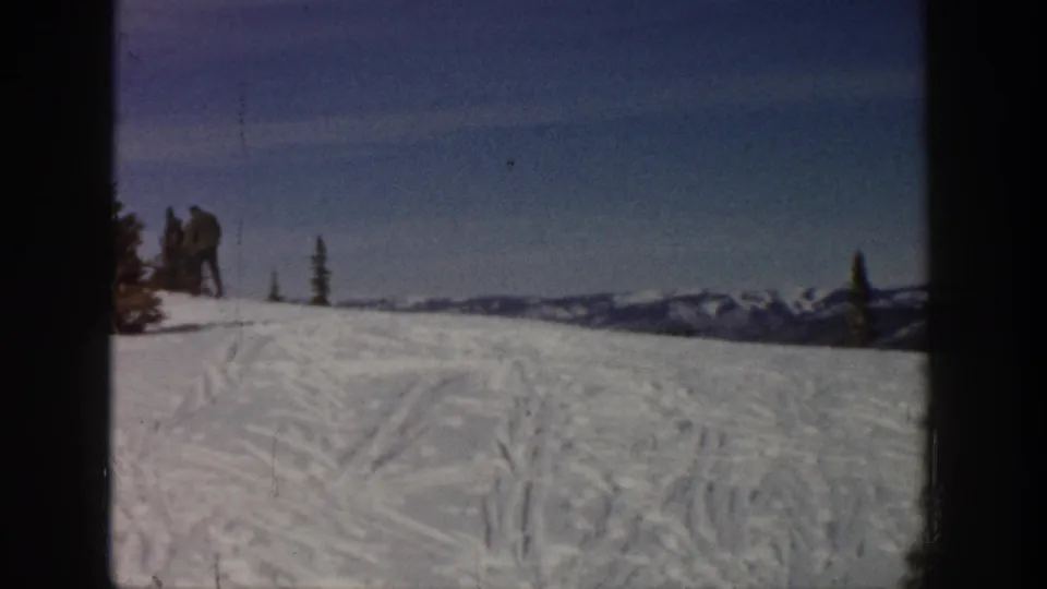 Distant skiers stand on a snowy Aspen ridge in 1961, with tracked snow, evergreens, and mountain peaks behind them.