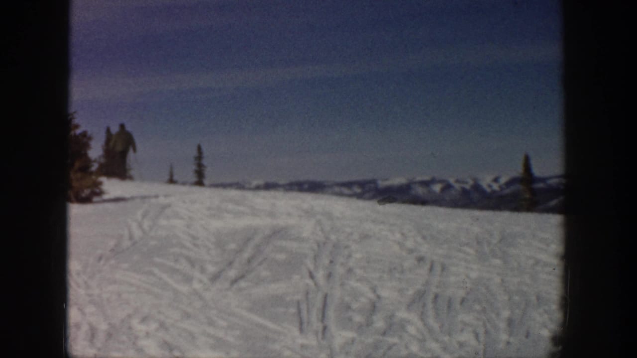 1961 Aspen home-movie frame showing a snowy slope with ski tracks, distant mountains, evergreen trees, and small skiers near the horizon.