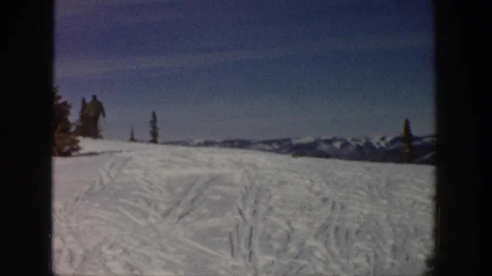 1961 Aspen home-movie frame showing a snowy slope with ski tracks, distant mountains, evergreen trees, and small skiers near the horizon.