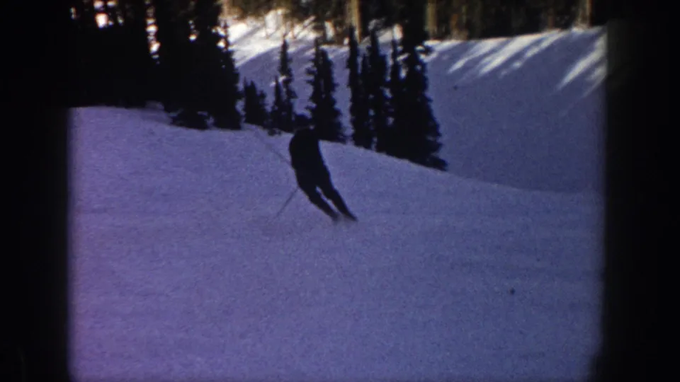 1961 Aspen home-movie frame showing a skier descending a snow-covered slope with evergreen trees and long shadows nearby.