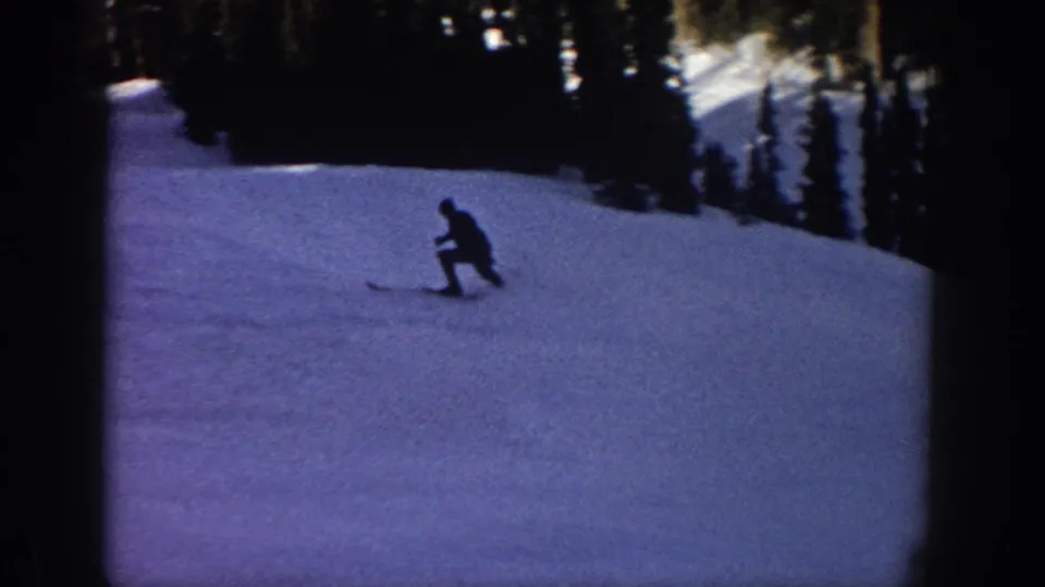 1961 Aspen ski-vacation frame showing a small skier crouched or turning on a snowy slope below evergreen trees.