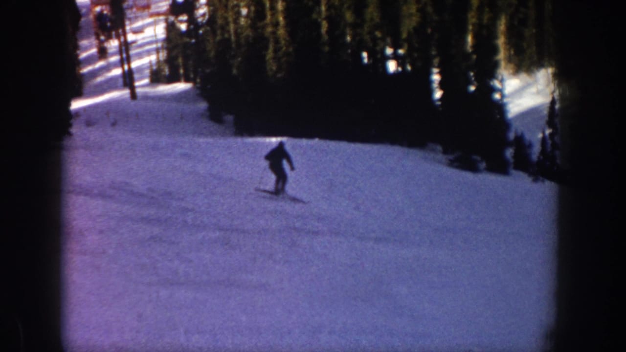 1961 Aspen home-movie frame of a small silhouetted skier crossing an open snowy run below evergreens.