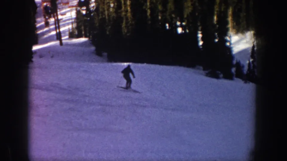1961 Aspen home-movie frame of a small silhouetted skier crossing an open snowy run below evergreens.