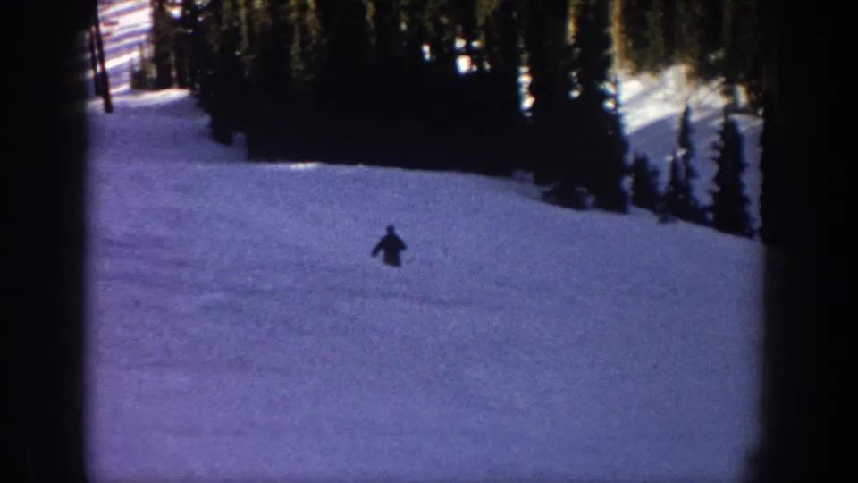 1961 Aspen 8mm frame showing a small dark skier lower on the snowy slope, framed by evergreens and soft film edges.