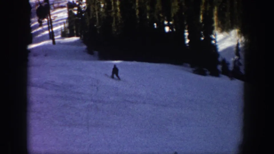 1961 Aspen home-movie frame with a silhouetted skier on a snow-covered run and dark evergreens along the upper slope.