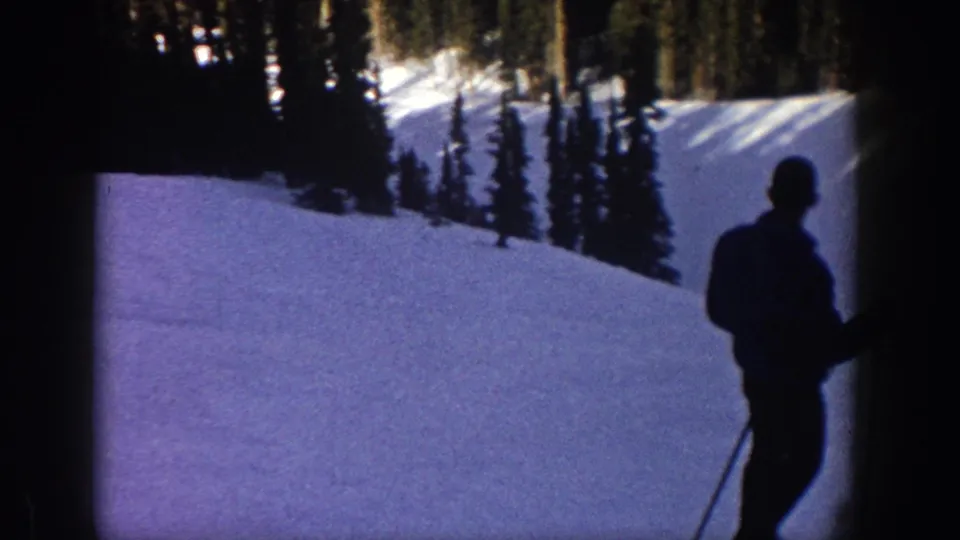 A foreground skier or observer stands with poles while another skier moves through the Aspen snow slope in 1961 footage.
