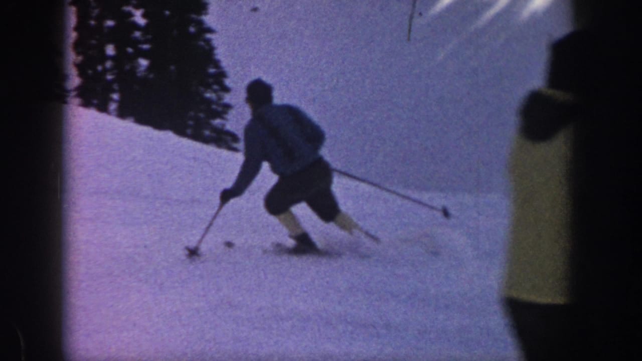 1961 Aspen home-movie frame showing a skier using poles while turning across a snowy slope bordered by dark evergreens.