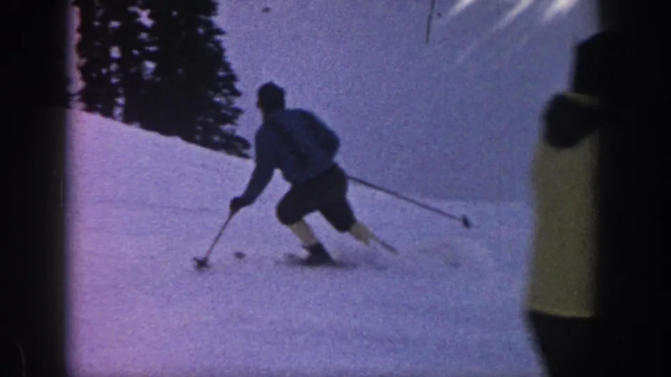 1961 Aspen home-movie frame showing a skier using poles while turning across a snowy slope bordered by dark evergreens.