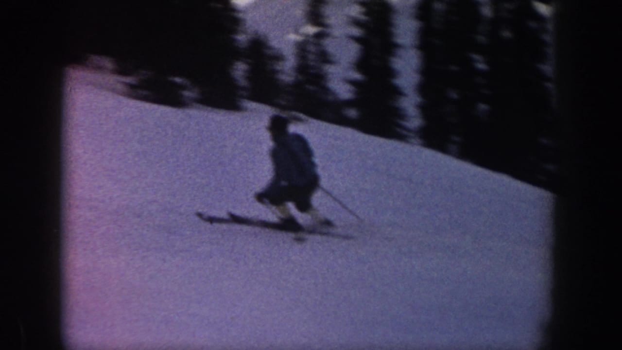 1961 Aspen footage frame with a skier low in a turn on a snow-covered slope, seen in dim blue-purple 8mm color.