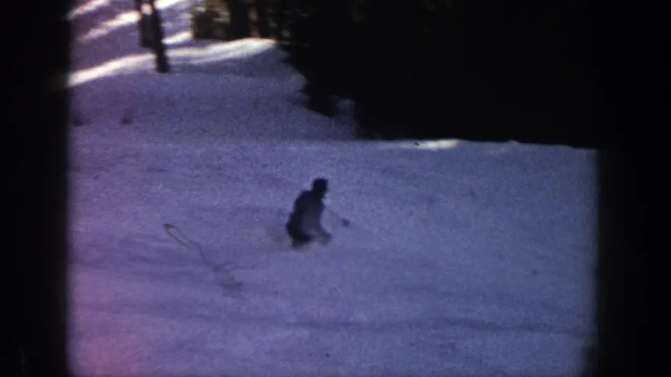 1961 Aspen frame showing a distant skier on a snowy slope, framed by dark trees and deep shadow in low-light 8mm footage.