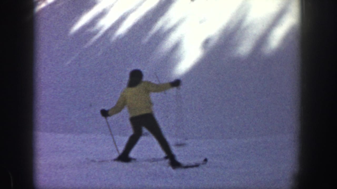 1961 Aspen home-movie frame showing a skier in a yellow jacket crossing a snowy slope with dark film borders.