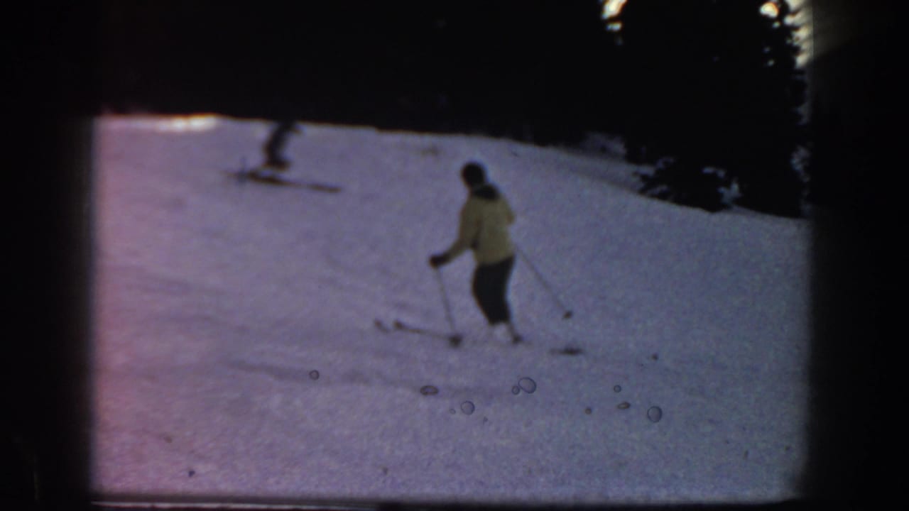 1961 Aspen home-movie frame showing a distant skier in a yellow jacket on a snowy slope near a dark tree line.