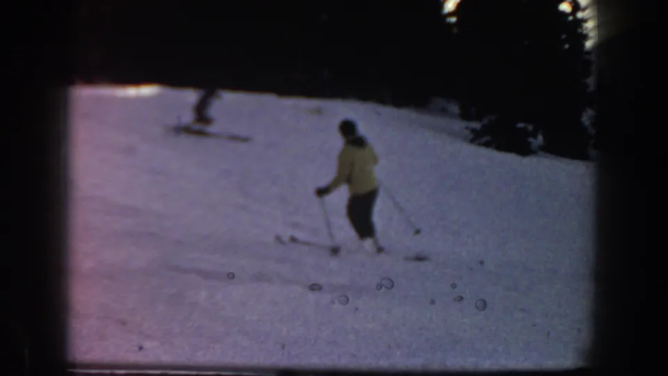 1961 Aspen home-movie frame showing a distant skier in a yellow jacket on a snowy slope near a dark tree line.