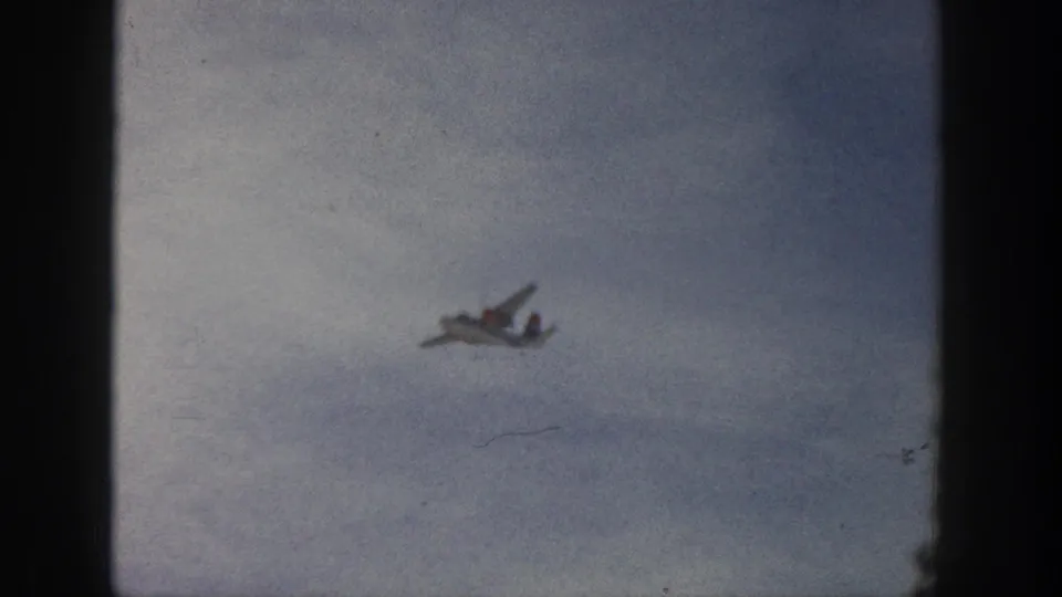 A distant small aircraft crosses a pale cloud field, framed by grain and dark 8mm gate edges.