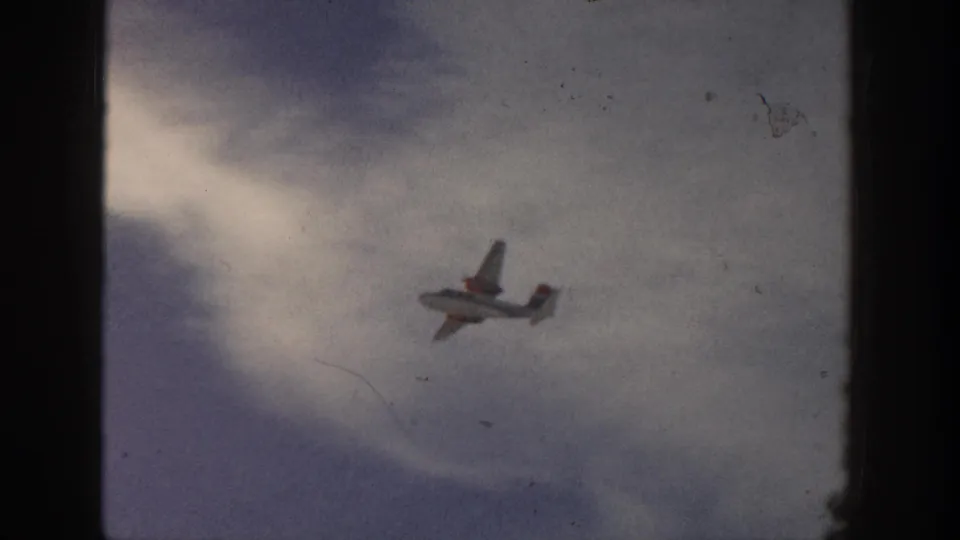 Small airplane shown closer against a clouded blue-gray sky, with visible mid-century 8mm film texture.