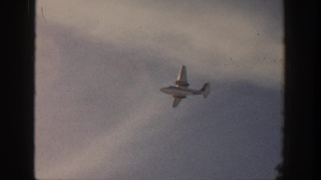 Small propeller airplane over Aspen, Colorado in 1961, seen against a pale clouded sky on faded 8mm film.
