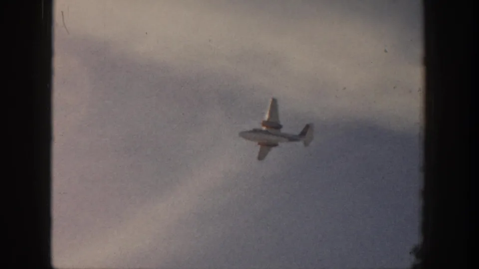 Small propeller airplane over Aspen, Colorado in 1961, seen against a pale clouded sky on faded 8mm film.