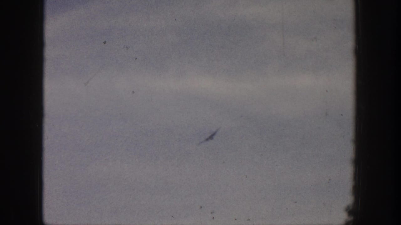 Faint small airplane silhouette against a pale cloudy sky with dark 8mm frame borders.