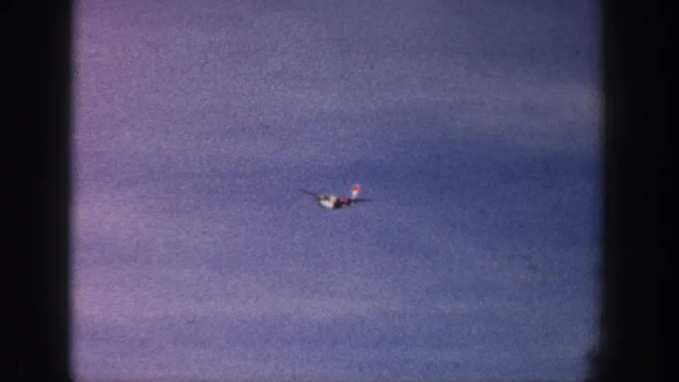 A small propeller airplane with red tail coloring is seen against a cloudy sky in supported 1961 Aspen footage.