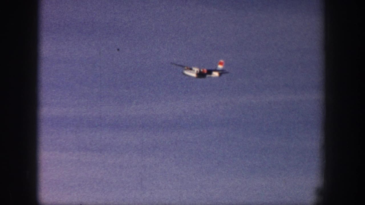 1961 Aspen home-movie frame showing a distant white single-engine airplane with red tail markings against a blue sky.