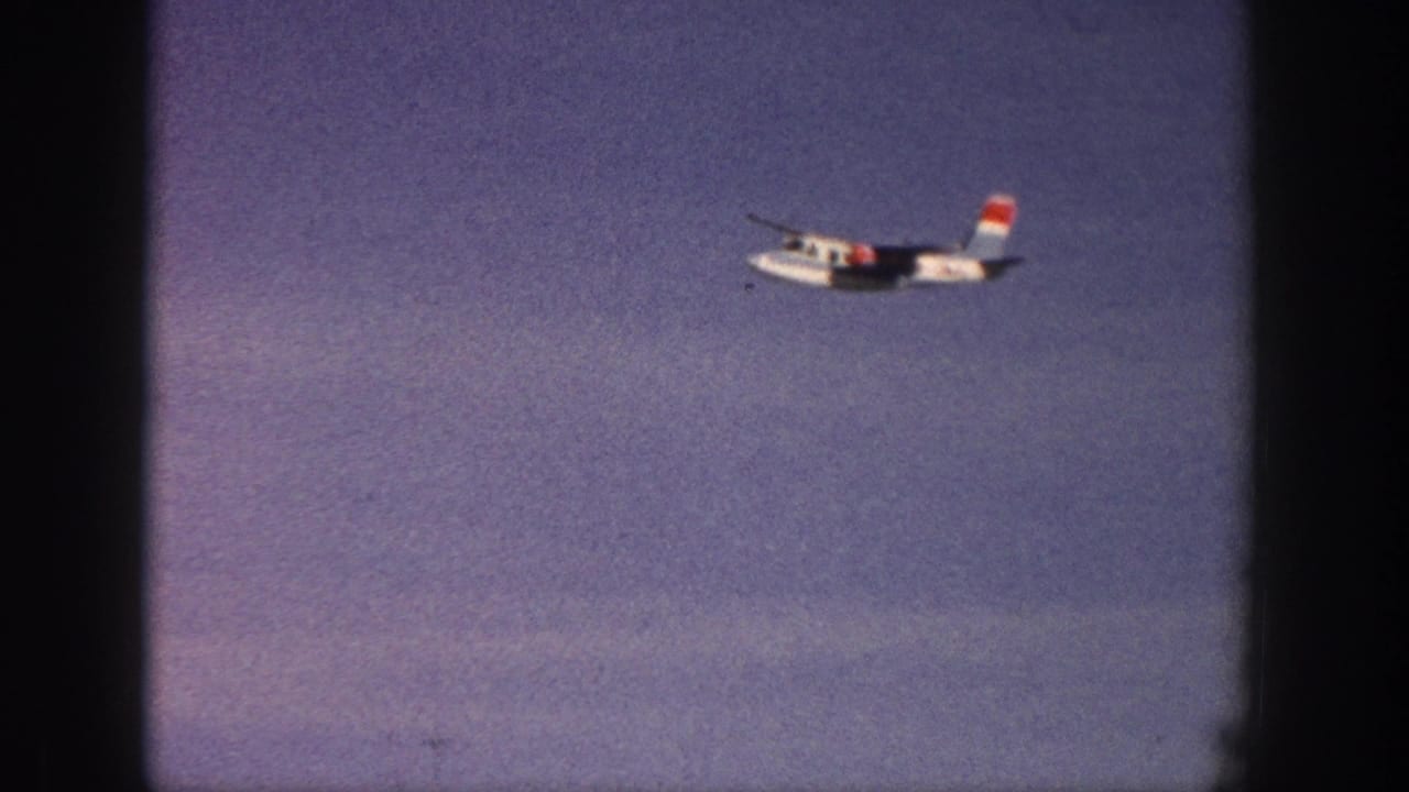 1961 Aspen 8mm frame with a small white propeller aircraft moving across a lightly clouded blue sky.