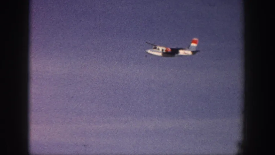 1961 Aspen 8mm frame with a small white propeller aircraft moving across a lightly clouded blue sky.
