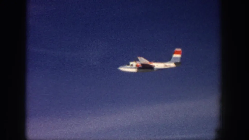 1961 Aspen home-movie frame showing a closer side view of a white propeller plane with red-and-blue tail markings.