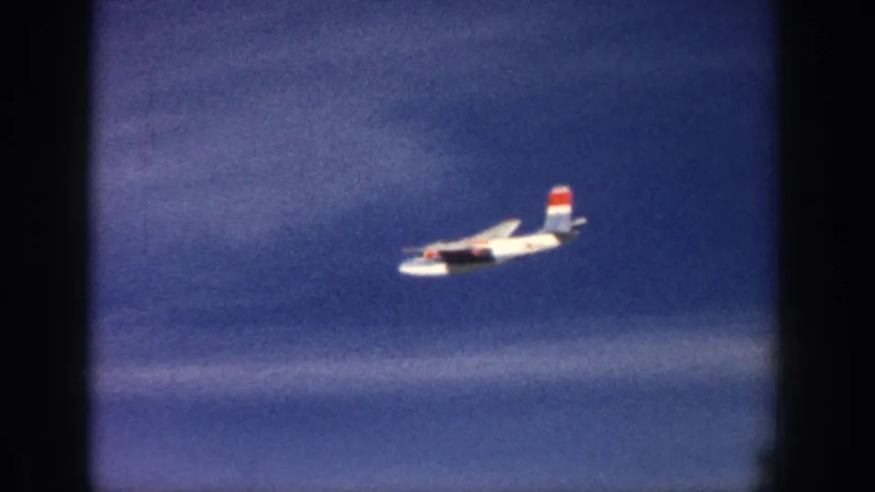 1961 Aspen 8mm frame of a white single-engine airplane moving rightward above a soft band of clouds.