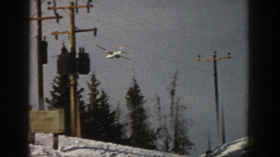 A low-flying propeller plane appears near the treeline, with evergreens, utility poles and snow visible below.