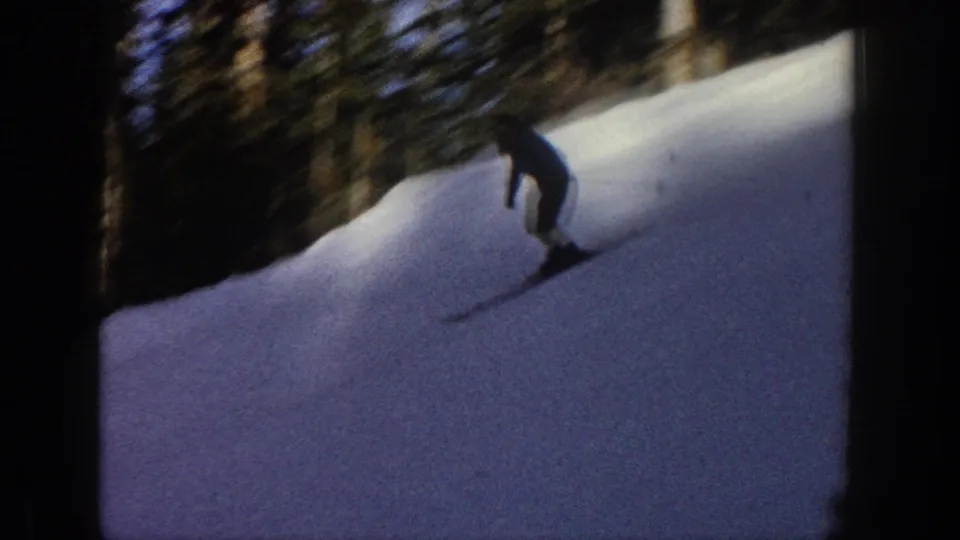 8mm frame of a skier leaning into a turn on a shaded Aspen snow slope beside evergreen trees.