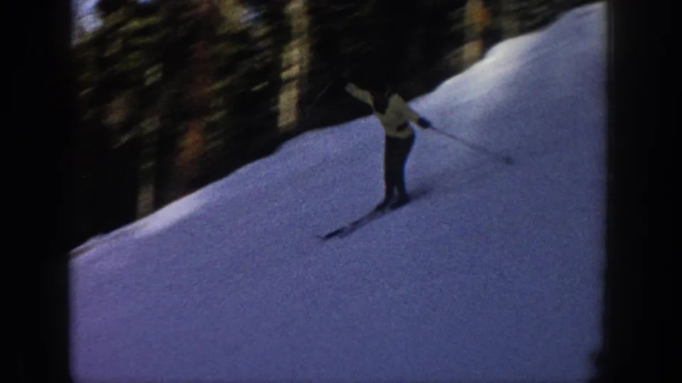 A skier in a light jacket leans into motion on a snowy wooded slope in 1961 Aspen home-movie footage.