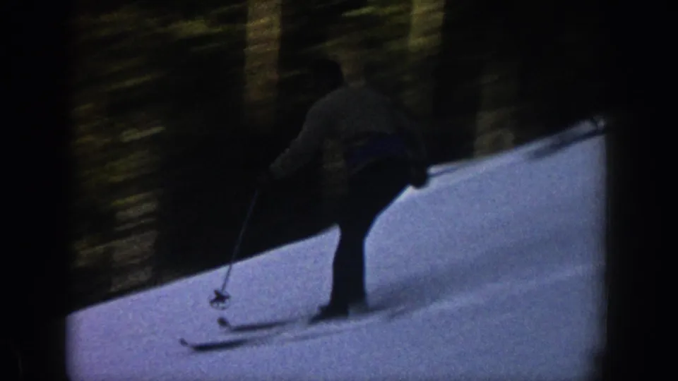 The skier passes closer to the camera on a shaded Aspen snow slope, framed by dark trees and 8mm film borders.