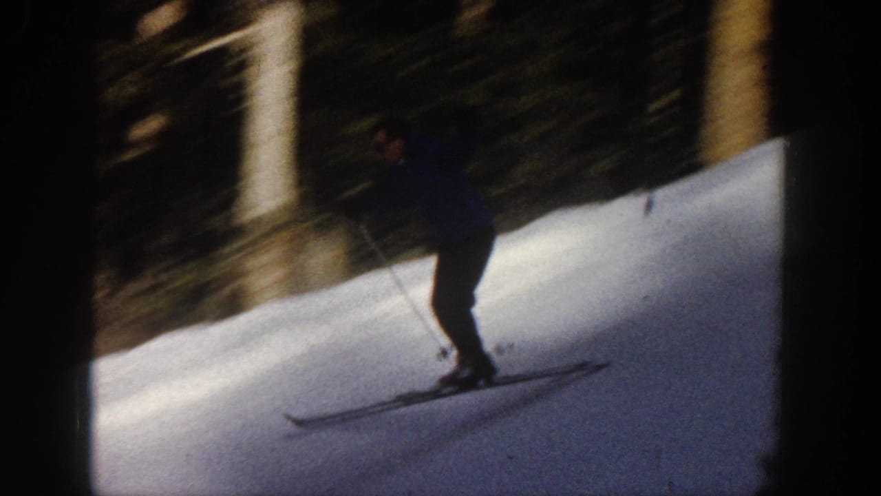 A skier descends a shaded snowy Aspen slope with poles and narrow skis, framed by dark trees in the 1961 home movie.
