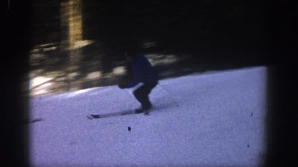 1961 Aspen 8mm frame with a skier crouched in a turn on snow, silhouetted against a dark wooded background.