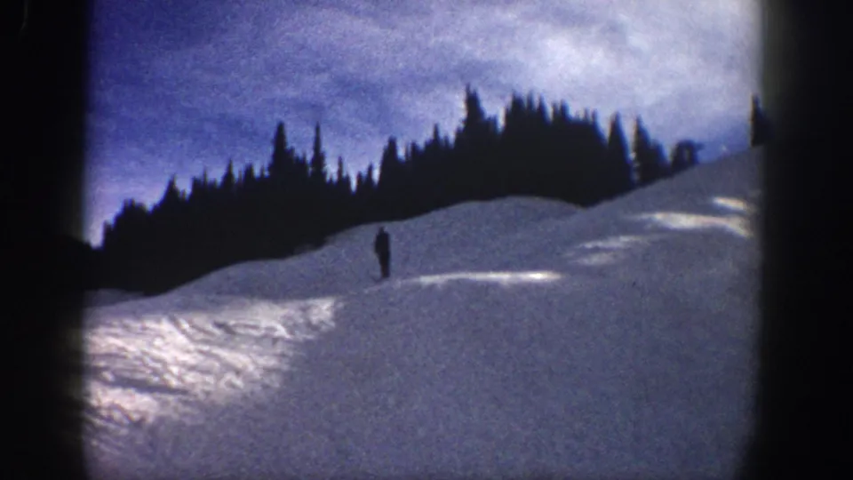 Wide 1961 Aspen home-movie view of a lone skier on a broad snow-covered hillside beneath evergreen trees.