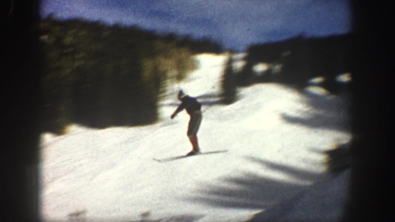 1961 Aspen home-movie frame of a distant skier moving across a sunlit snowy slope with evergreen trees above.